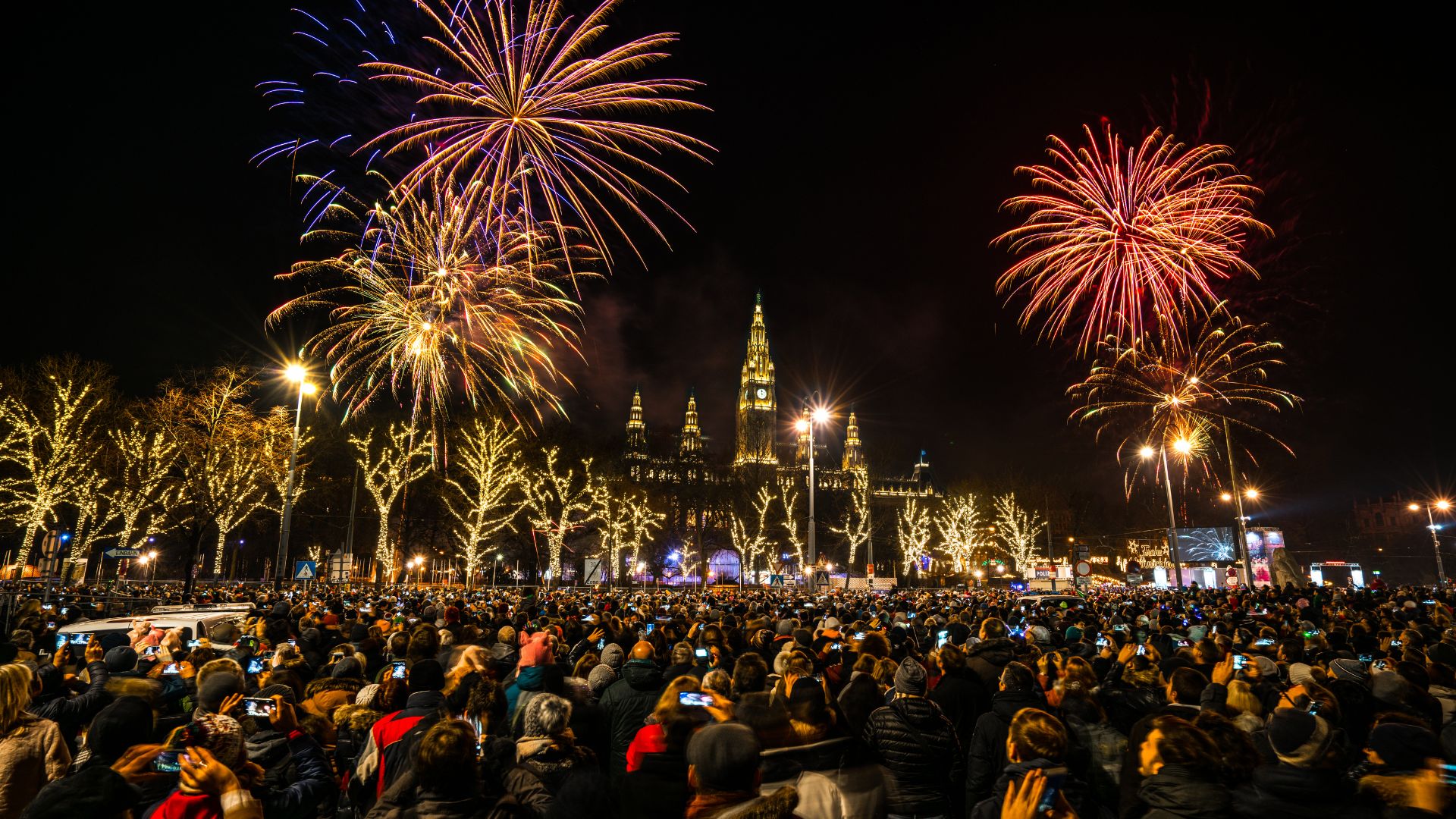 ALT描述: 歐洲風格建築物前的跨年盛況，密密麻麻的群眾舉起手機拍攝夜空中金紅色的巨大煙火。