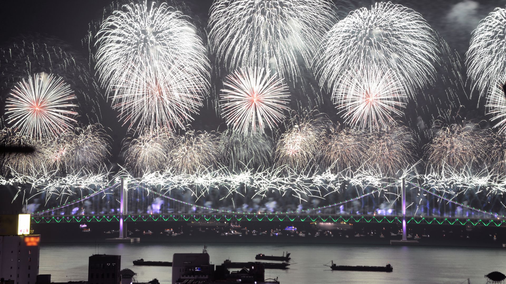 ALT描述: 廣闊的跨海大橋夜景，橋面上方排開多朵巨大的白色與紅色煙火，水面上停泊著數艘船隻。