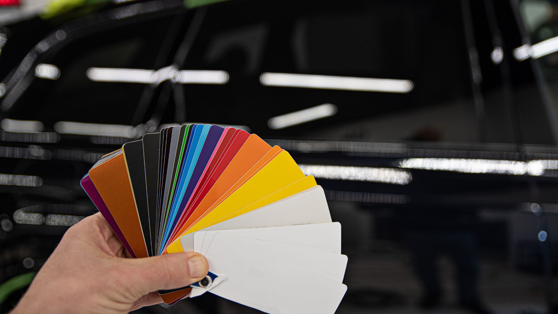ALT描述: 一隻手拿起一本扇形展開的多色車貼、彩繪膜色卡，背景是一輛擁有鏡面光澤的黑色汽車。 