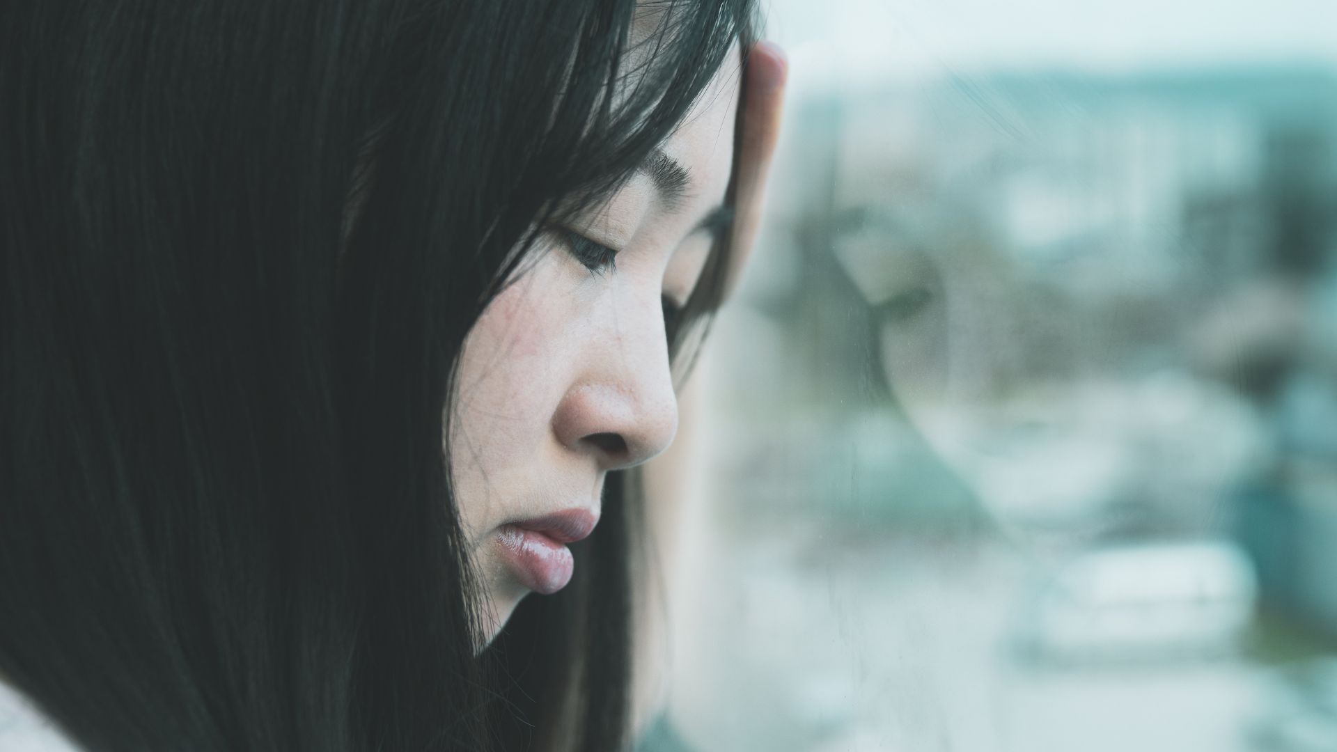 ALT描述: 一名年輕女子靠在窗邊，手扶著額頭，眼神憂鬱地凝視著窗外。