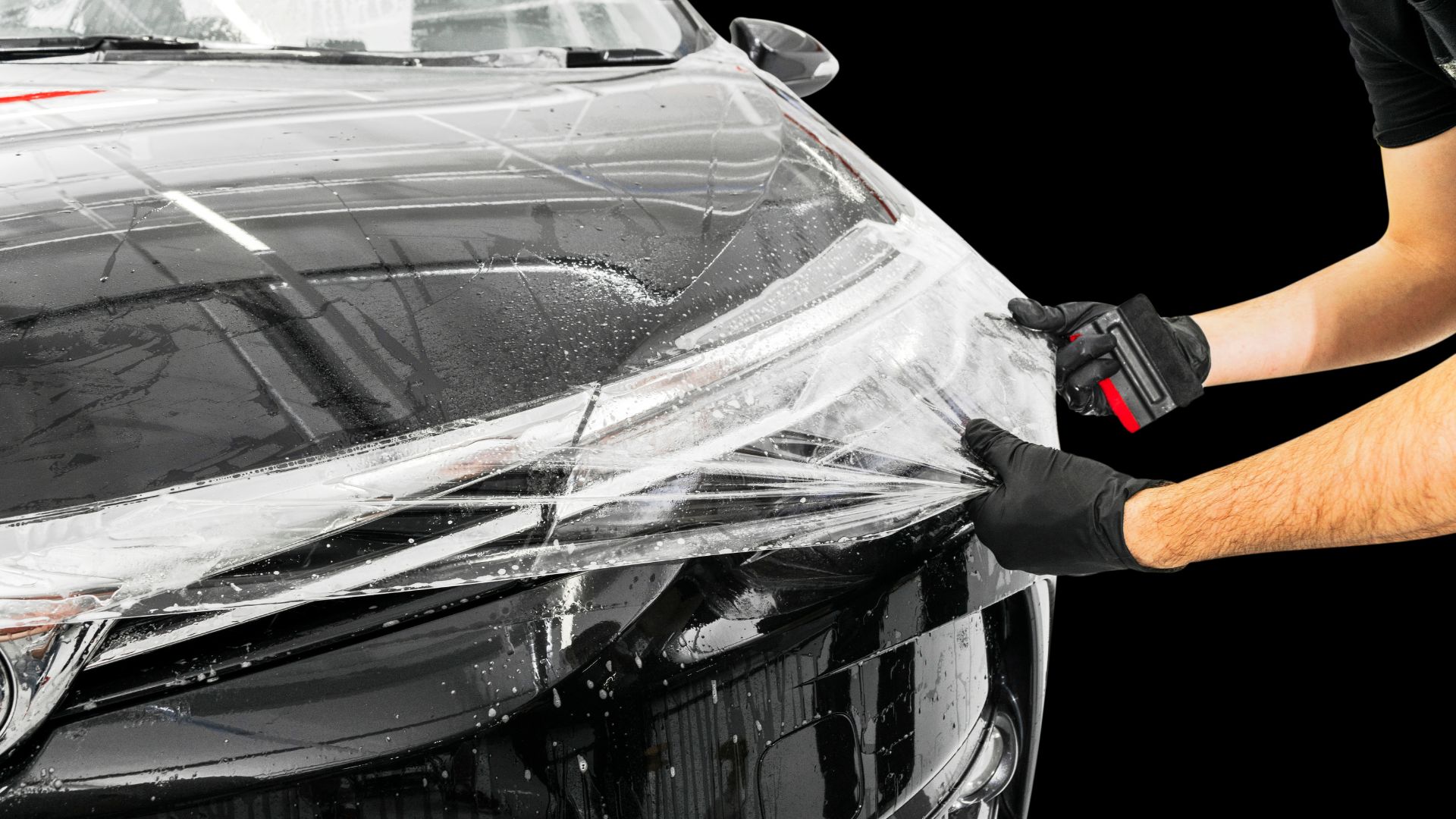ALT描述:一個人戴著黑色的手套，兩隻手正在調整汽車引擎蓋上的保護膜。右手拿著一個紅色的刮刀，用來去除膜下的氣泡。