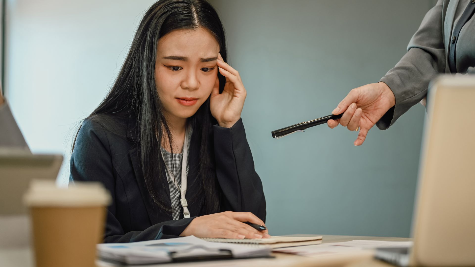 ALT描述: 在辦公室內，一名亞洲女子身穿黑色西裝，右手扶太陽穴，低頭看著桌上的筆記本和文件，表情焦慮。旁邊一名男子身穿灰色西裝，右手拿著一支黑色筆遞向女子。桌上放著咖啡杯和文件，背景為藍灰色牆面。