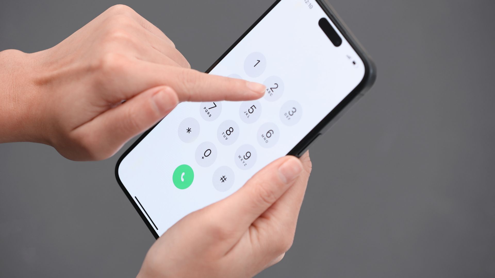 ALT描述: 近距離特寫雙手拿著智慧型手機，手指正準備點擊螢幕上的數字撥號介面，背景為簡潔的灰色調。