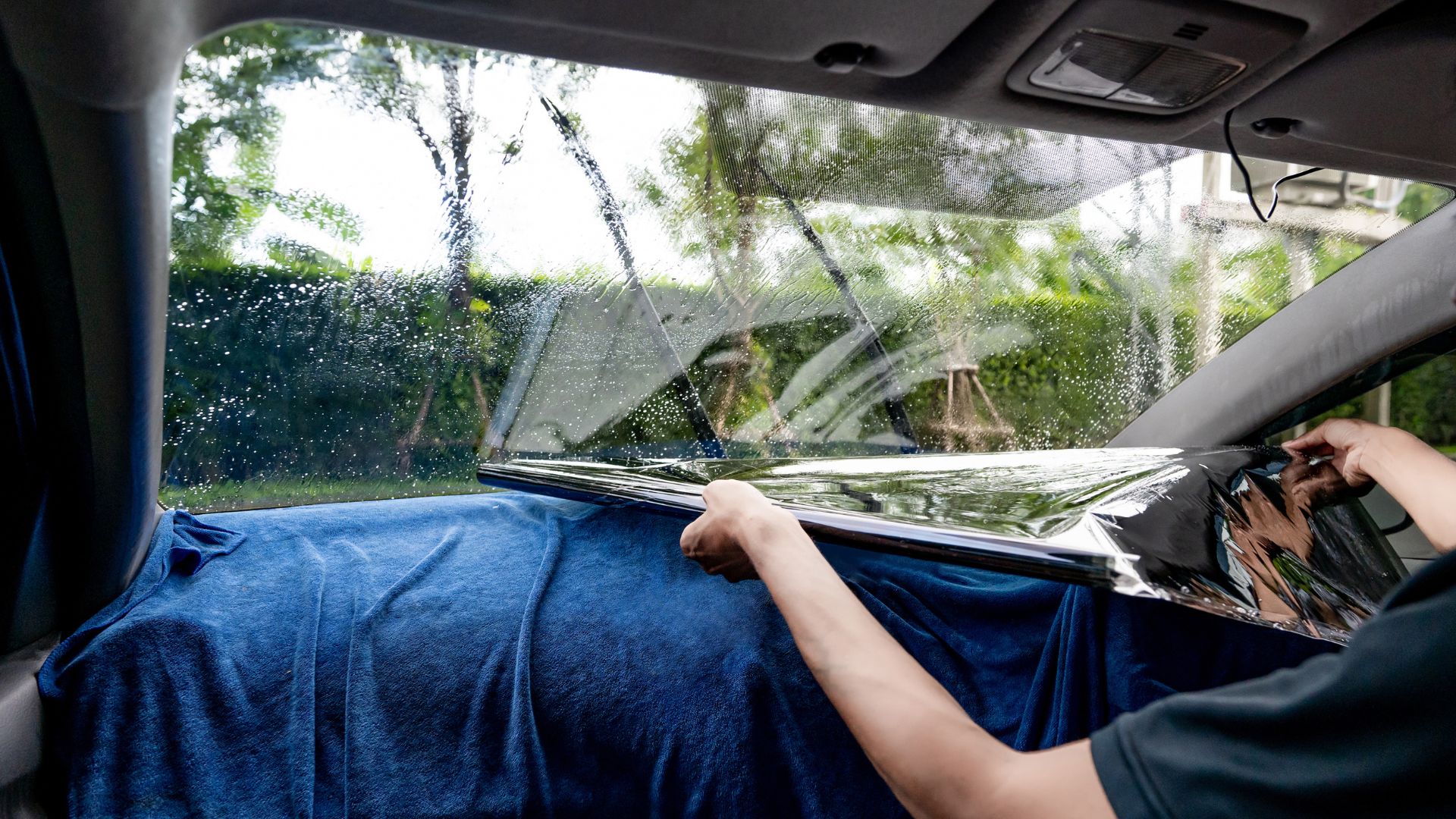 ALT描述: 從車內視角看，安裝隔熱紙的技術人員正將一張裁切好的隔熱紙移向已噴水的擋風玻璃，儀表板上舖著藍色毛巾防止水滴落下。
