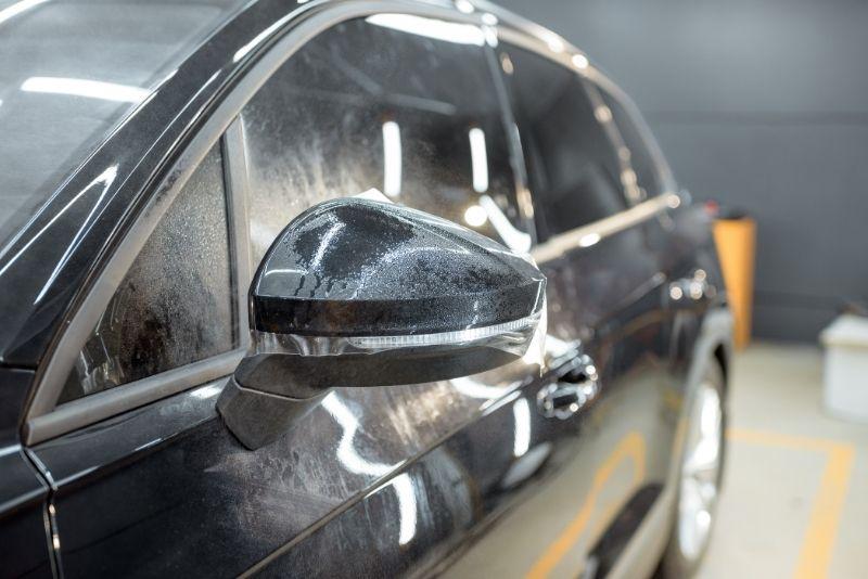 ALT描述: 專業汽車美容工作室內，一輛黑色汽車的後視鏡和車窗上貼著剛噴過水的透明車漆保護膜（PPF），表面佈滿細密水珠，反射出工作室的明亮燈光。