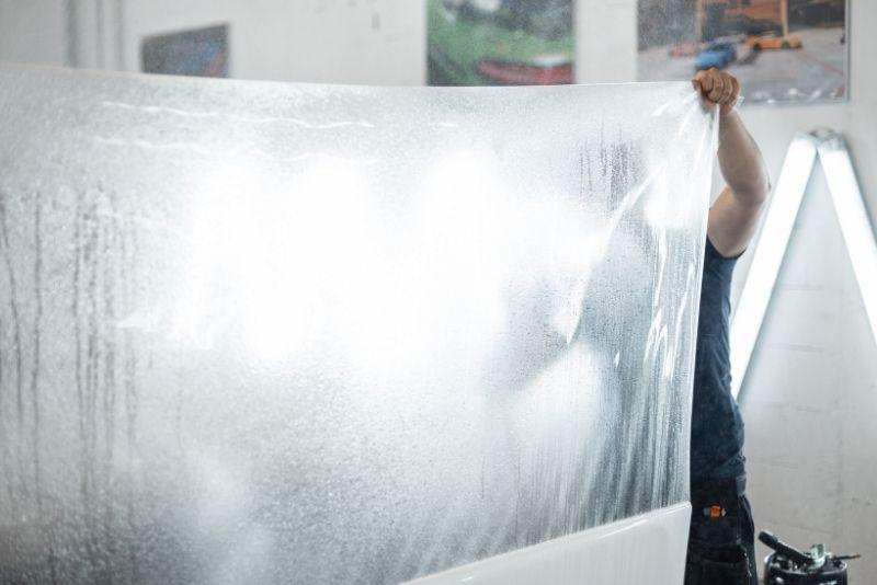 ALT描述: 汽車貼膜技師在霧氣繚繞的工作室內，雙手撐開一張巨大的、表面佈滿水珠的透明犀牛皮保護膜，準備將其貼在下方的白色汽車上。