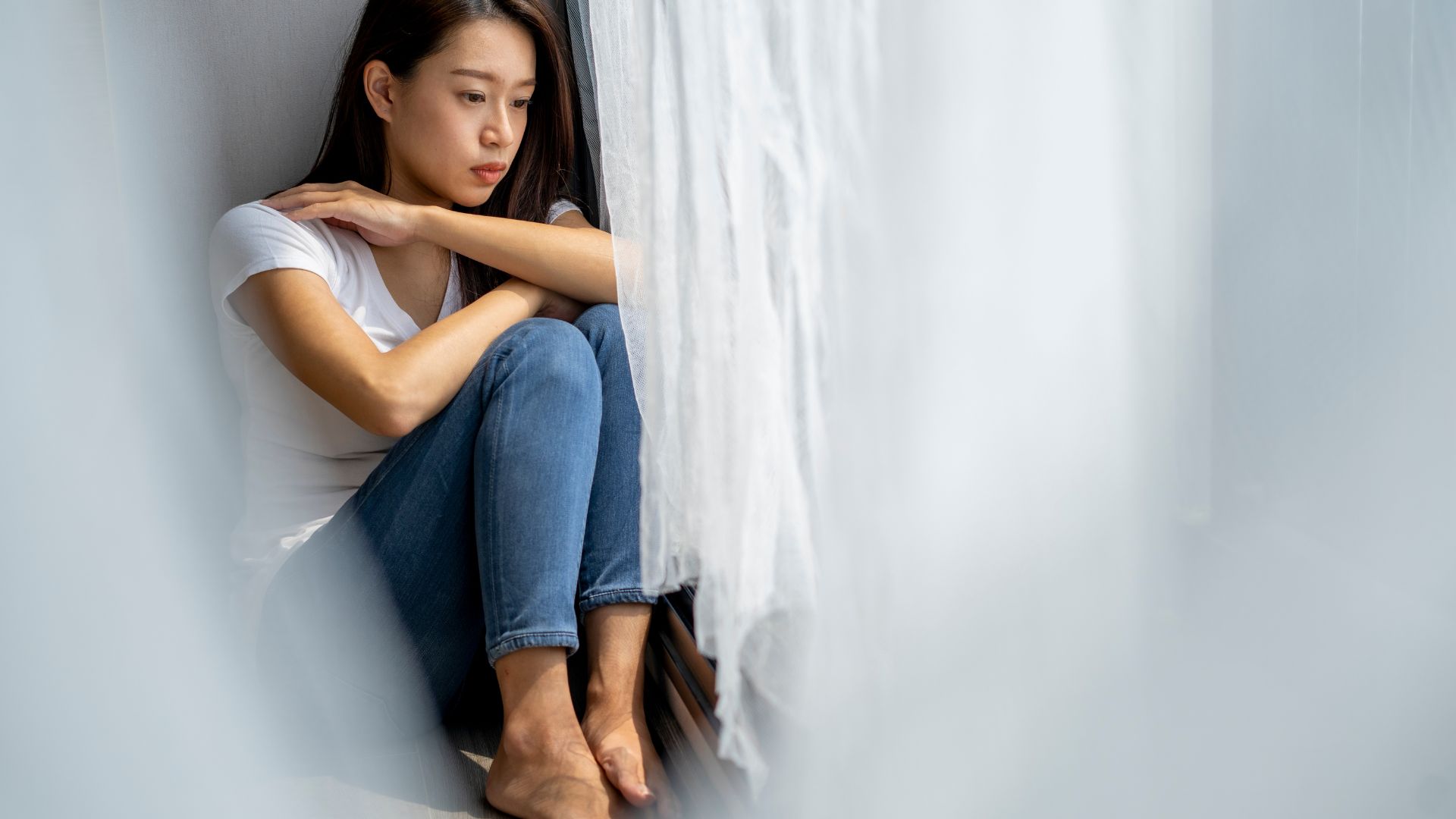 ALT描述: 一名穿著白色 T 恤與牛仔褲的女性獨自坐在窗邊的地板上，雙手環抱膝蓋，眼神憂鬱地望向窗外，流露出孤獨或悲傷的氛圍。