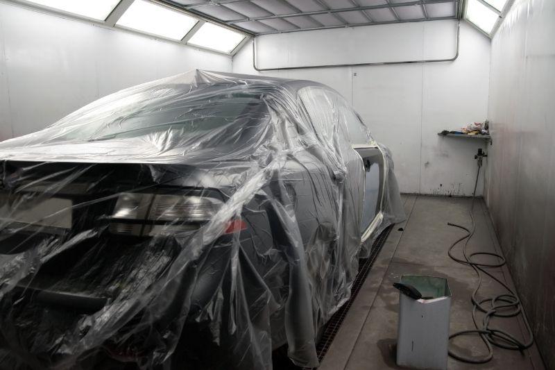 ALT描述: 在專業的汽車噴漆房內，一輛深灰色的轎車被大面積的透明塑膠薄膜完全覆蓋，準備進行噴漆或施工，地面上散落著桶子和電線。