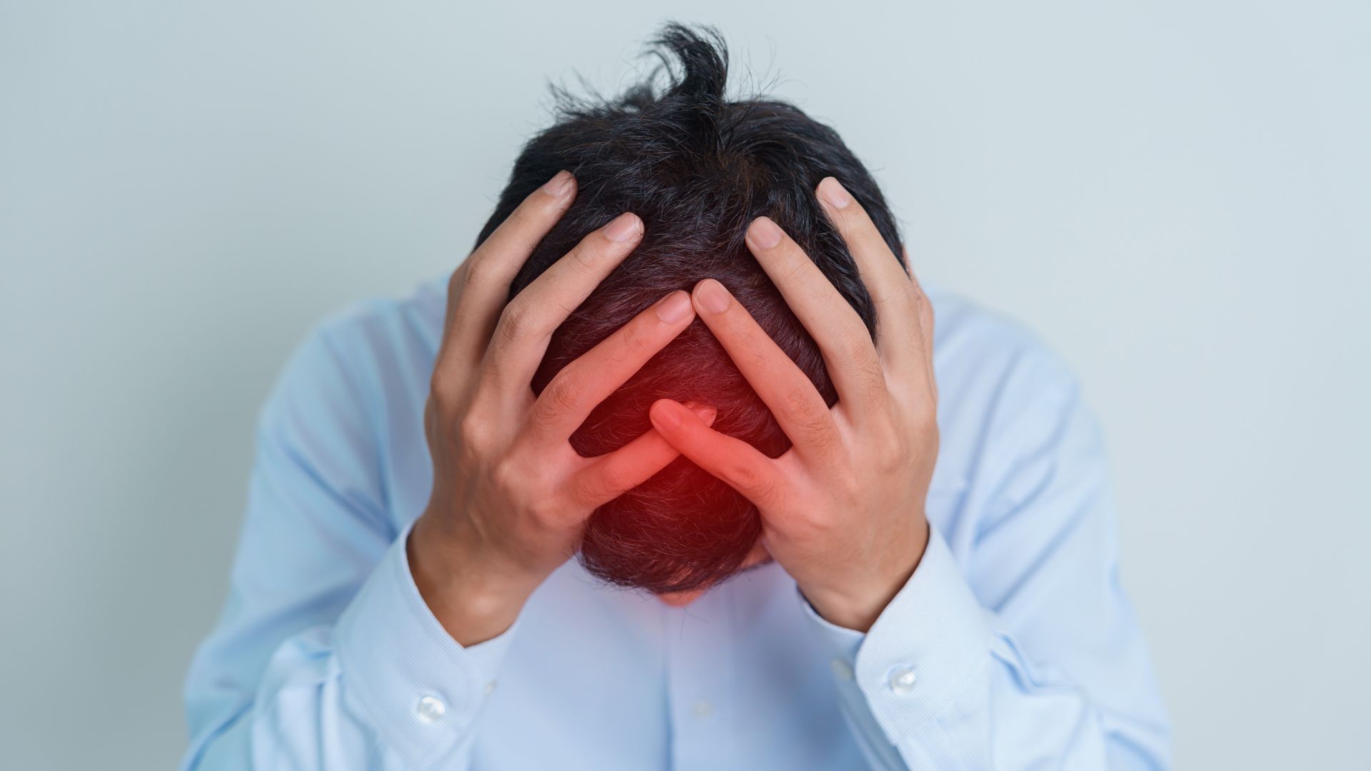 ALT描述: 一名亞洲男子身穿淺藍色襯衫，雙手抱頭，手指交叉，低頭坐著。頭部頂端有紅色高亮區域，暗示疼痛或不適。背景為淡灰藍色牆面。