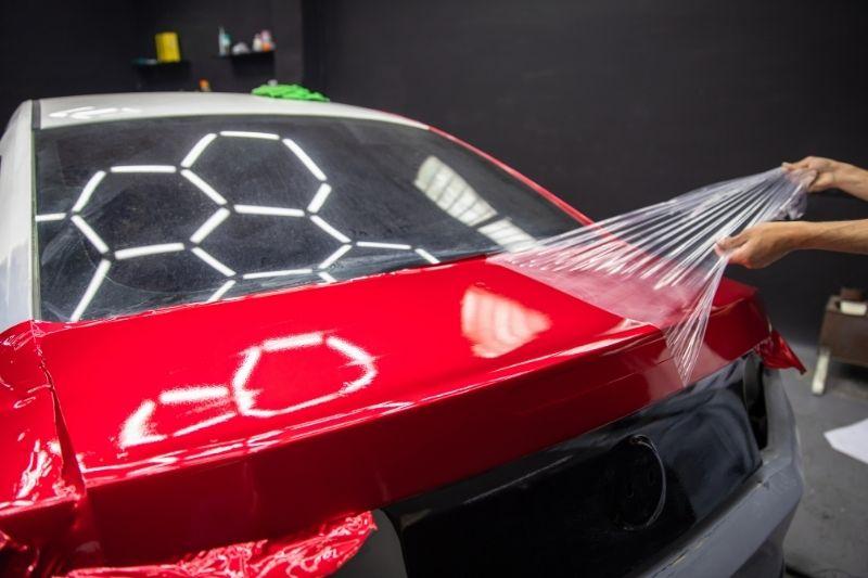 ALT描述: 在汽車包膜工作室中，一名技師正在處理一輛剛貼好鮮紅色包膜的白色轎車尾部，他正雙手拉扯著行李箱蓋邊緣多餘的透明保護膜。