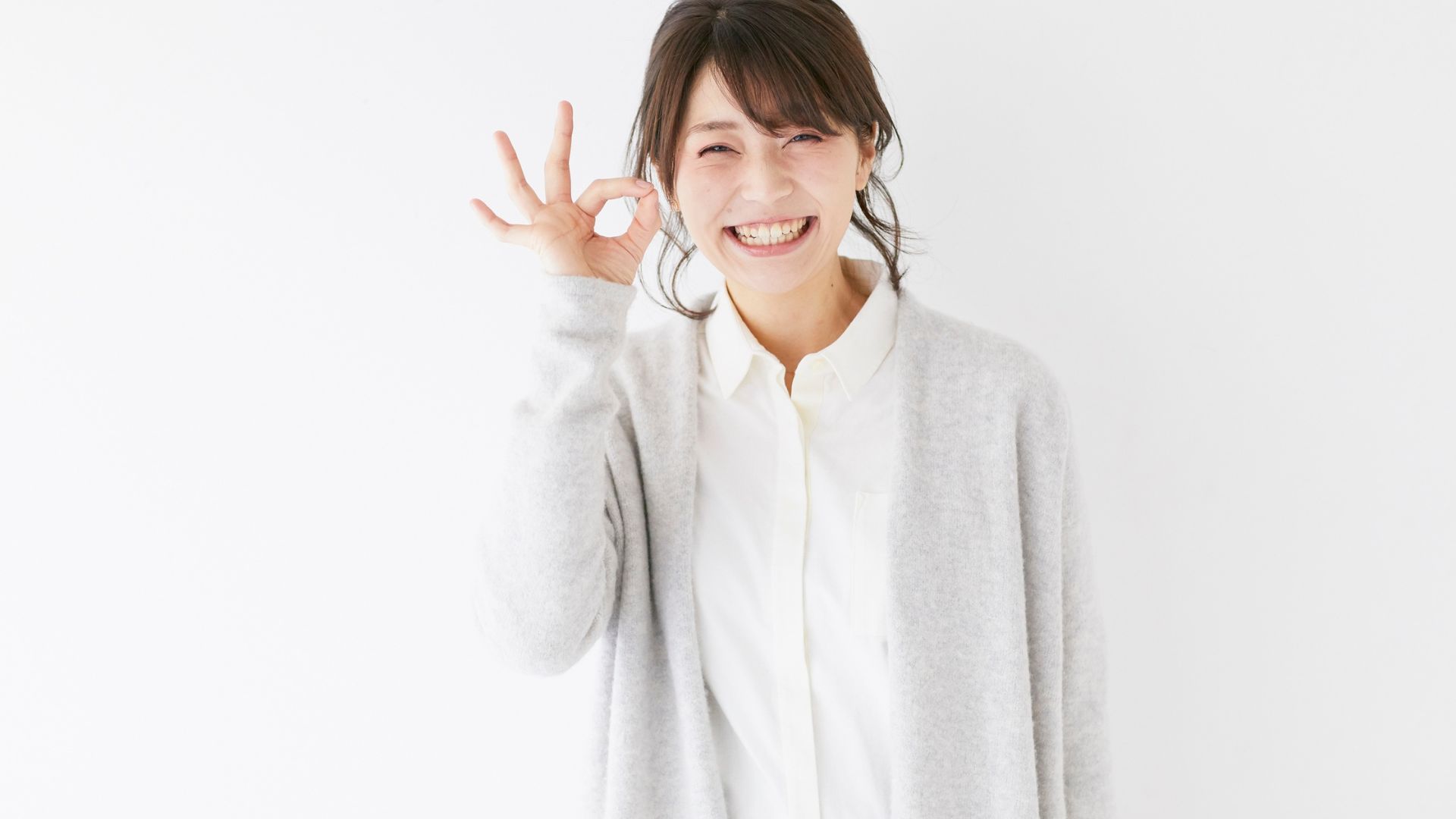 ALT描述: 一名女子身穿白色鈕扣長袖襯衫和淺灰色針織外套，對著鏡頭燦爛微笑，並用右手做出「OK」的手勢。