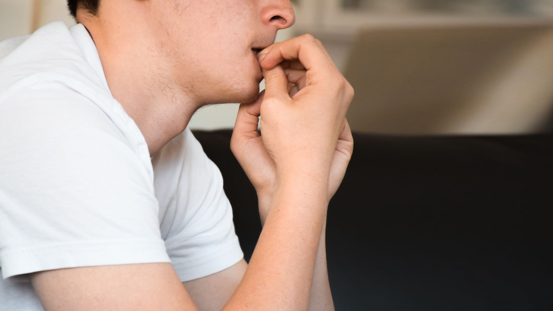 ALT描述: 一名年輕男子坐在黑暗的沙發上，身穿白色圓領短袖上衣，雙手交叉捂住嘴巴，眼神空洞，表情焦慮。