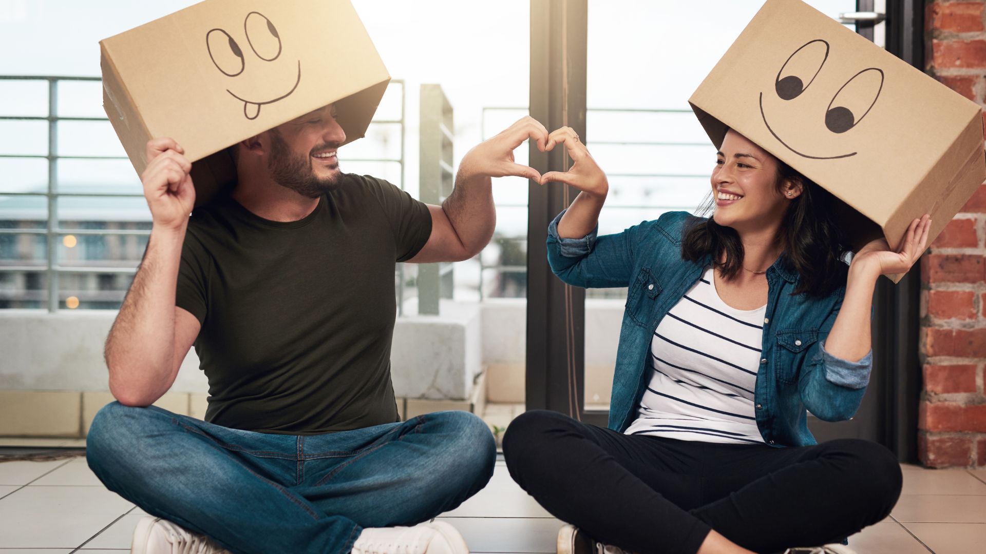 ALT描述: 一對男女坐在地板上，頭上各套著一個畫有大笑表情的紙箱，兩人用手共同比出一個愛心形狀，氣氛俏皮且溫馨。