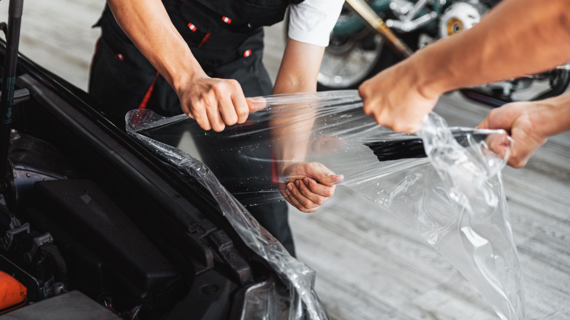 ALT描述: 技術人員正合力將透明的犀牛皮（汽車保護膜）拉伸、貼合在一輛黑色的汽車車頭蓋上。